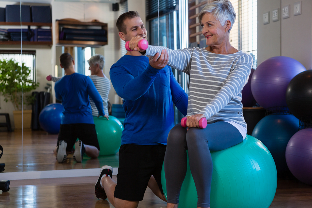 Senior woman on exercise ball, working out with assistance of male personal trainer